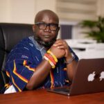 Ken Ashigbey, convener of the Media Coalition Against Galamsey, seated confidently at his office desk wearing traditional Ghanaian attire and bracelets, symbolizing his relentless advocacy against illegal mining in Ghana.