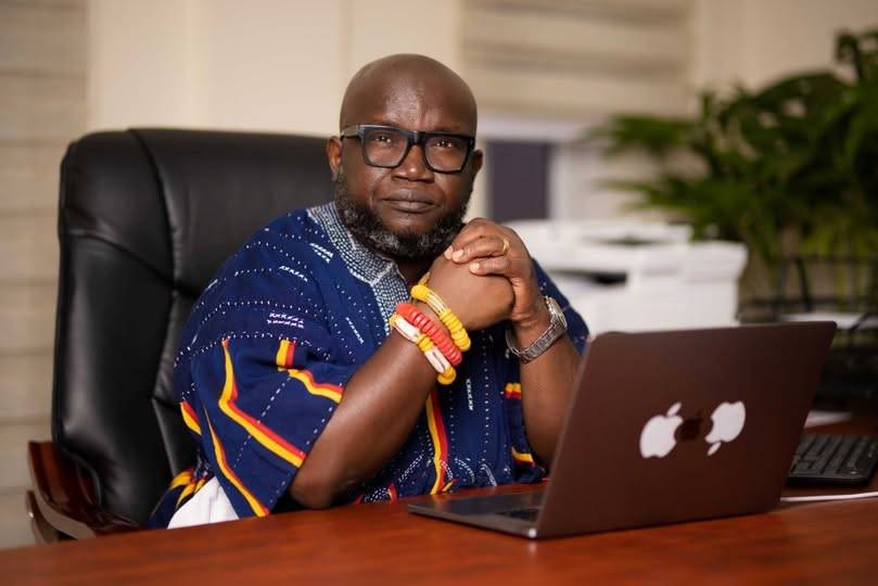 Ken Ashigbey, convener of the Media Coalition Against Galamsey, seated confidently at his office desk wearing traditional Ghanaian attire and bracelets, symbolizing his relentless advocacy against illegal mining in Ghana.